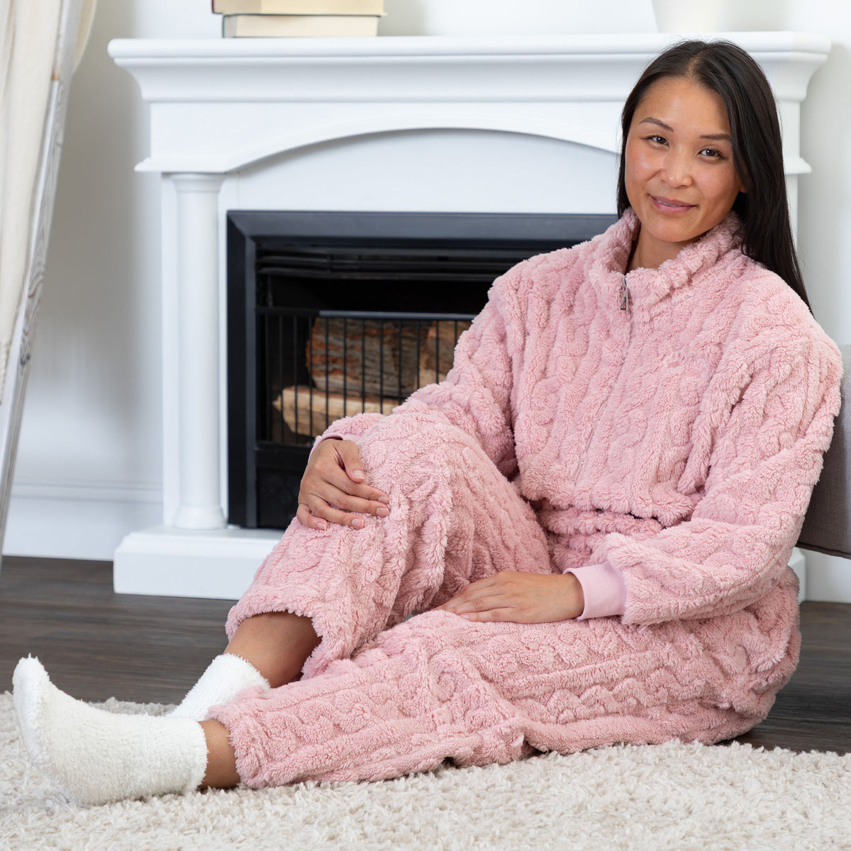 A woman wearing a cozy pink cable-knit fleece pajama set sits on a white rug in front of a white fireplace. She is wearing white socks.