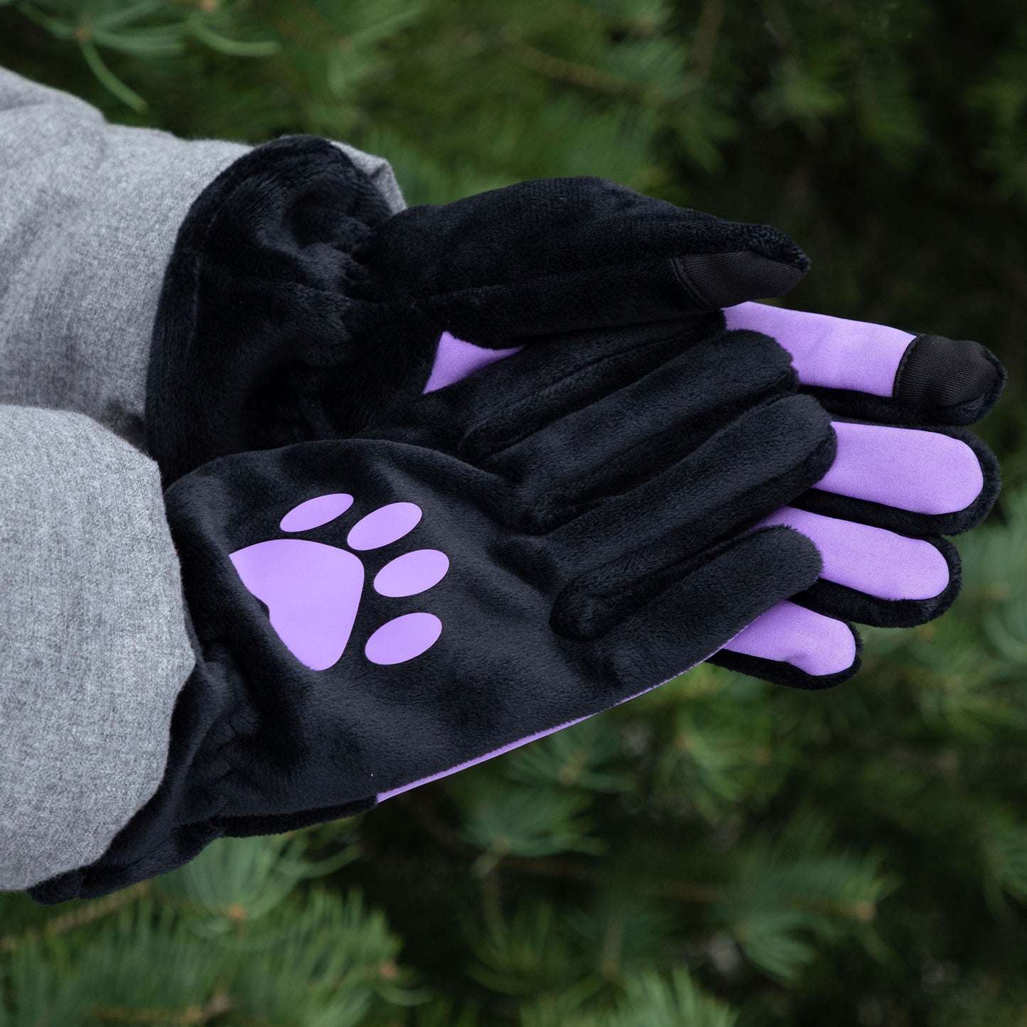 A person wearing black fleece gloves with purple paw prints against a green outdoor background.