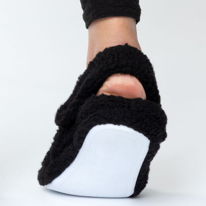 Black fuzzy slipper with white sole worn on a foot against a light gray background