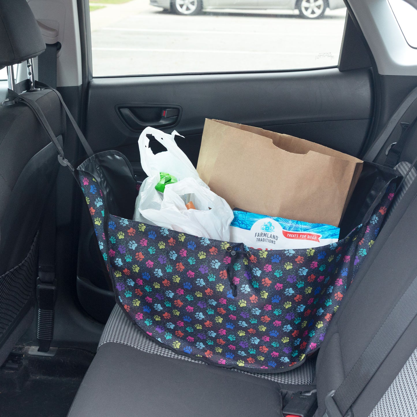 A floral car seat organizer bag filled with grocery items, including a paper bag, plastic bags, and a box of Farmers Supply toilet paper, sitting on a car's back seat.