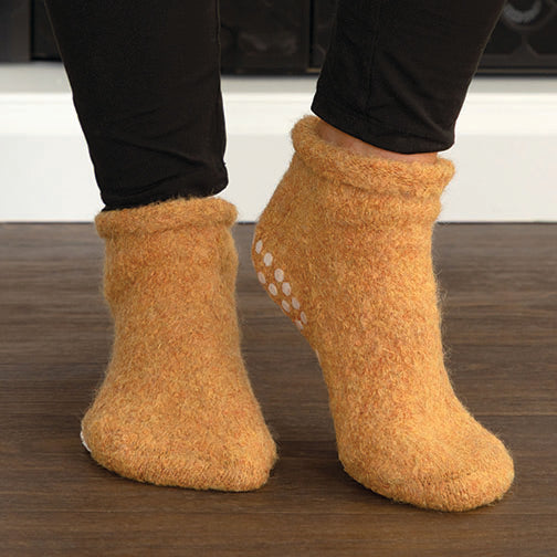 A pair of fuzzy ankle socks in a warm mustard yellow-orange color, worn with black leggings on a dark wood floor. The socks have white grip dots on the sole.