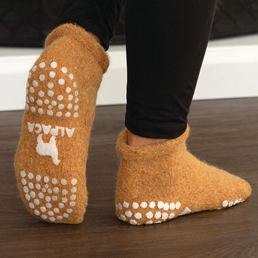 A person wearing fuzzy orange slipper socks with white grip dots and a white ALPACA logo on the sole, standing on a dark wood floor.