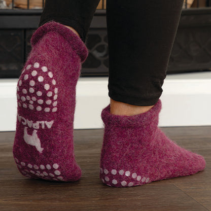 A person wearing purple felted slippers with white grip dots and a white alpaca graphic on the sole, standing on a dark wood floor.