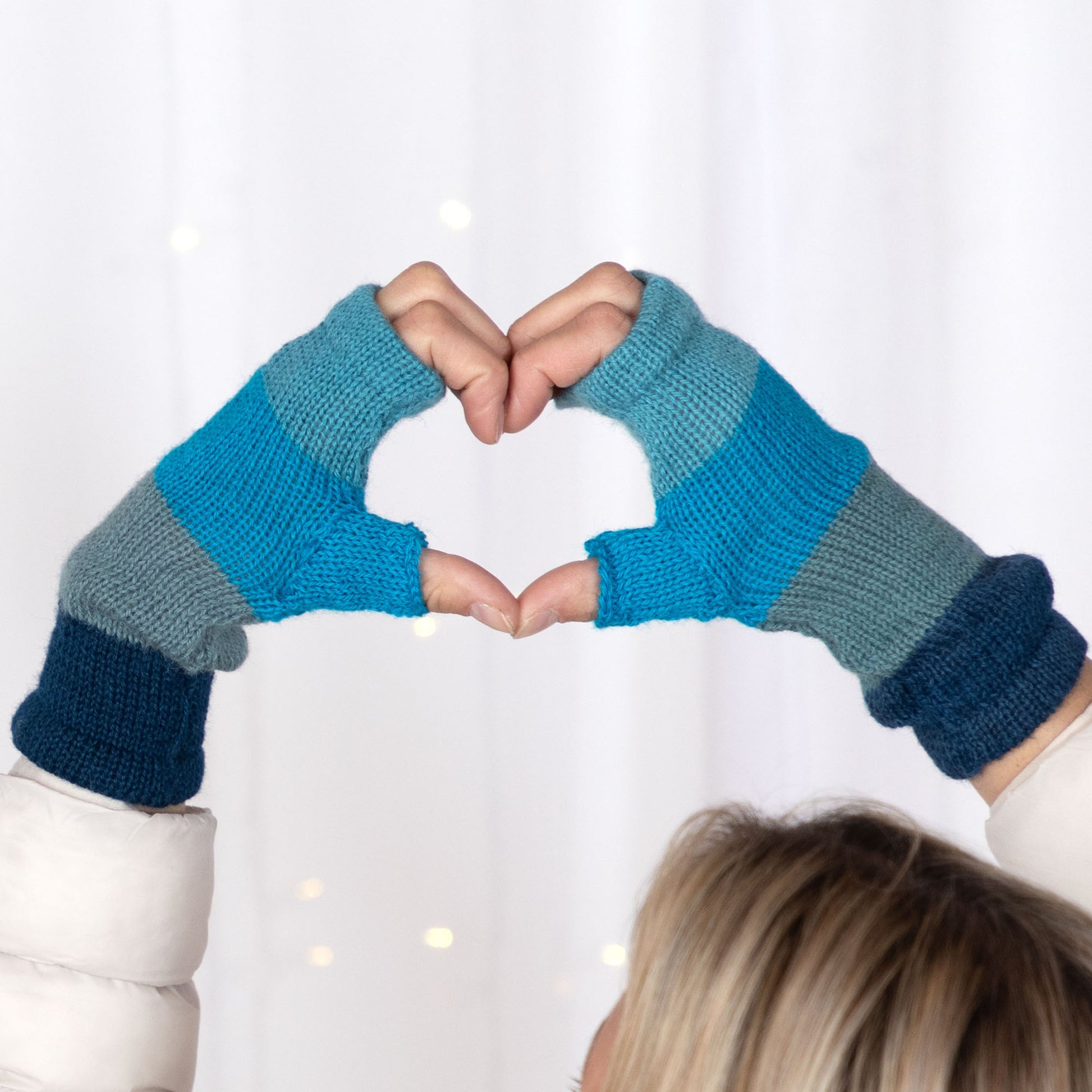 Person wearing blue and gray striped fingerless gloves making a heart shape with their hands on a white background.