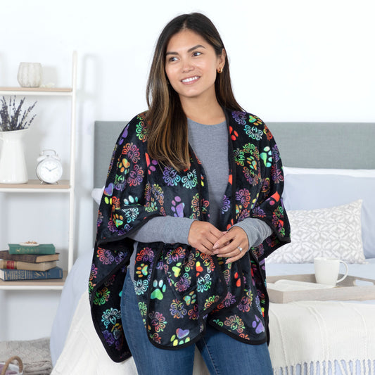 A woman standing in a cozy bedroom. She is wearing a black based fleece, pocketed shawl, patterned with orange, green, purple and cyan paw prints and decadent designs. The edges are lined with black piping.