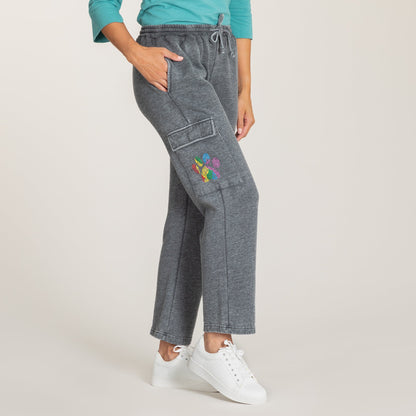 A person modeling gray heather cargo jogger pants with a colorful embroidered patch on the left thigh, paired with a teal top and white sneakers against a light beige studio background.