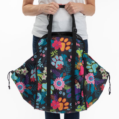 A person holding a large black duffel bag with a colorful floral and paw-print pattern, shown against a plain white background.
