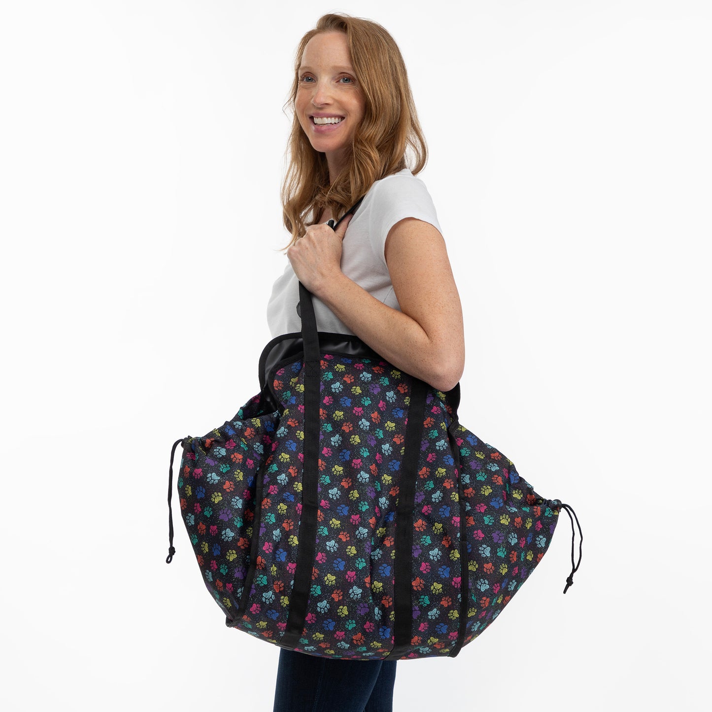 A woman wearing a white t-shirt and dark jeans carries a large black drawstring duffel bag with a colorful floral pattern, set against a plain white studio background.