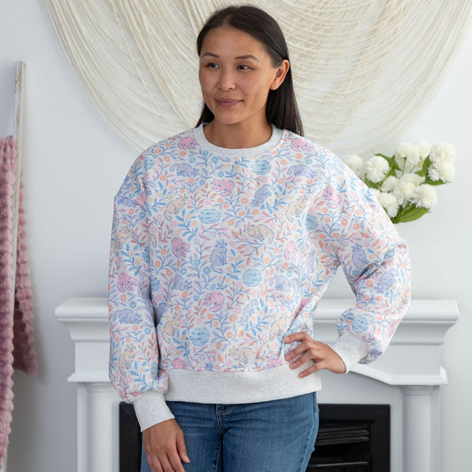 A woman wearing a long-sleeve crewneck sweatshirt with a pastel floral pattern in pink, blue, and purple on a white background, paired with blue jeans, standing in front of a white fireplace mantel.