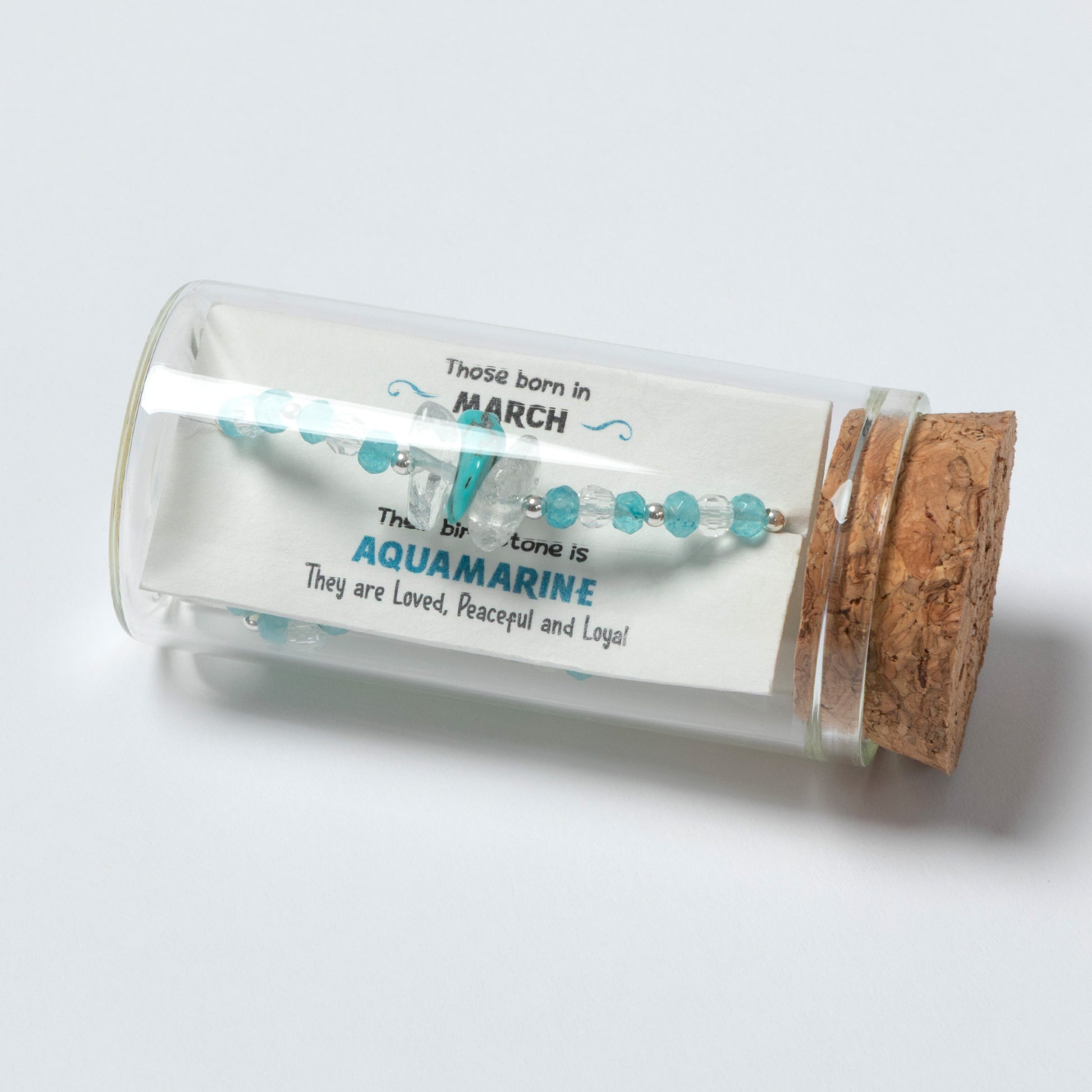 Glass jar with cork lid containing a bracelet labeled for March birthstone Aquamarine on a light gray background.