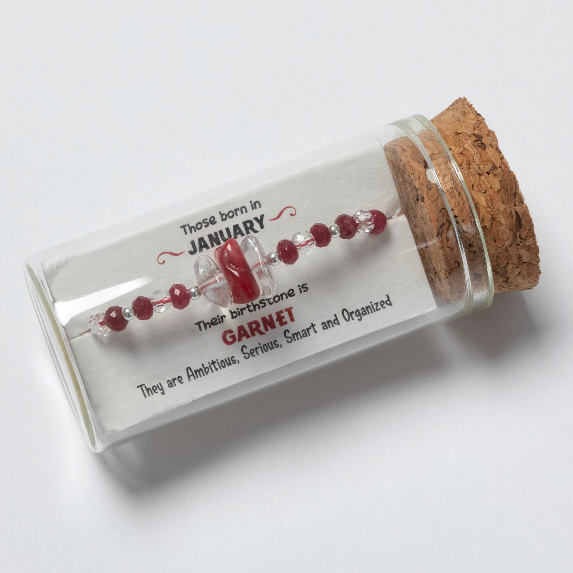 Glass bottle with cork lid containing a red gemstone and a label about January birthstones on a white background.