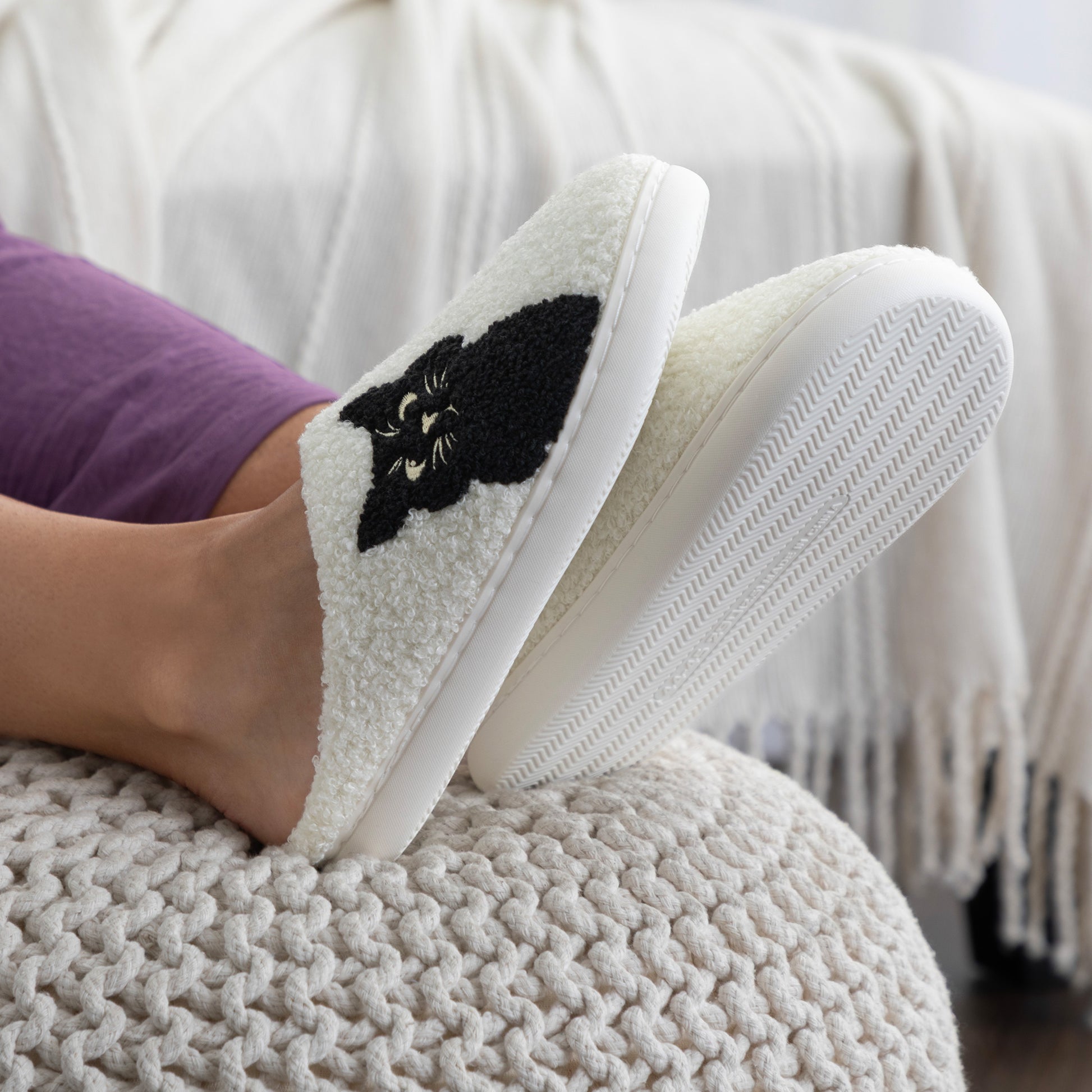 White slippers with black cat design worn by a person sitting on a textured surface.