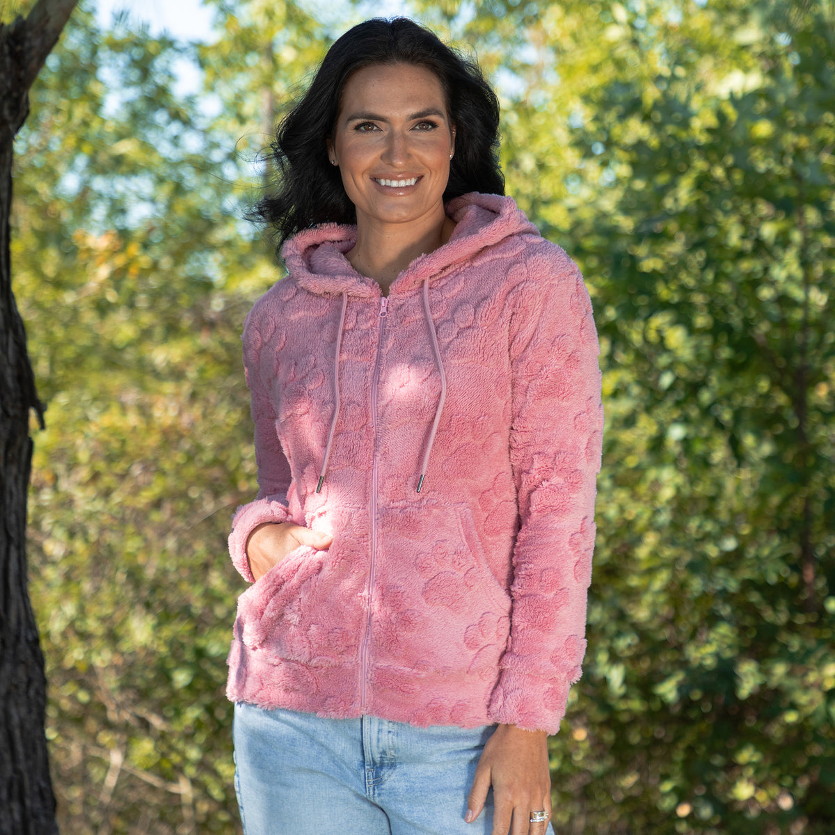 A woman wearing a pink textured fleece zip-up hoodie with a drawstring hood and front pockets, paired with light blue jeans, standing outdoors in front of a blurred green forest background.