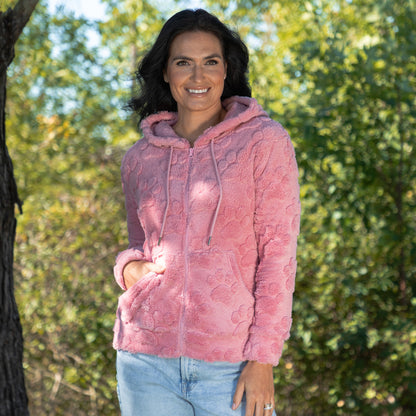 A woman wearing a pink textured fleece zip-up hoodie with a drawstring hood and front pockets, paired with light blue jeans, standing outdoors in front of a blurred green forest background.