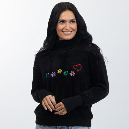 A woman wearing a black fuzzy turtleneck sweater with colorful embroidered paw prints and a red heart on the chest, photographed against a light gray studio background.
