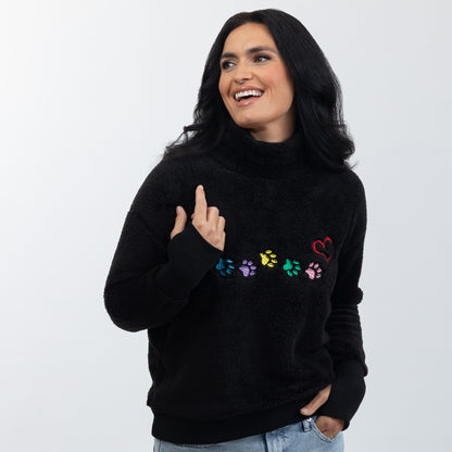A woman wearing a black fuzzy turtleneck sweater with colorful embroidered paw prints and a red heart on the chest, paired with light blue jeans, against a plain light gray background.