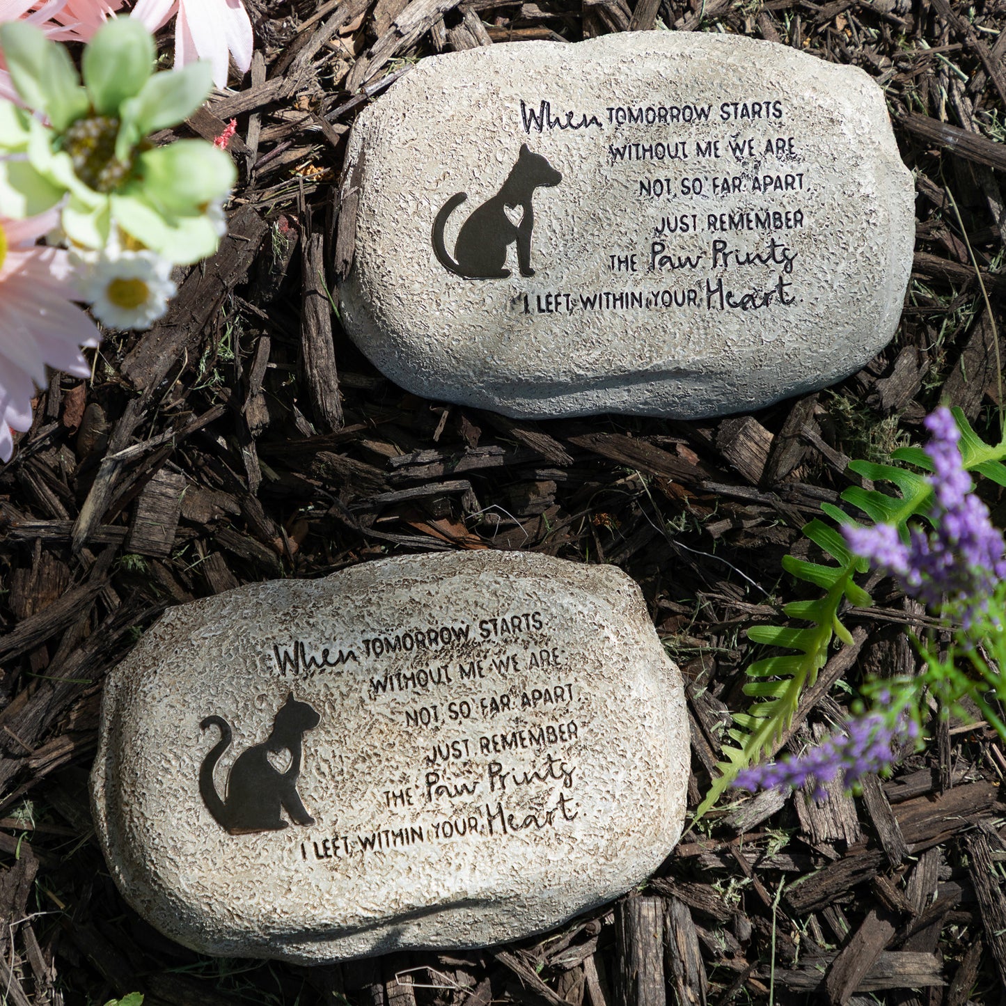 Two engraved stones with cat and dog illustrations and text on a mulched surface with flowers.