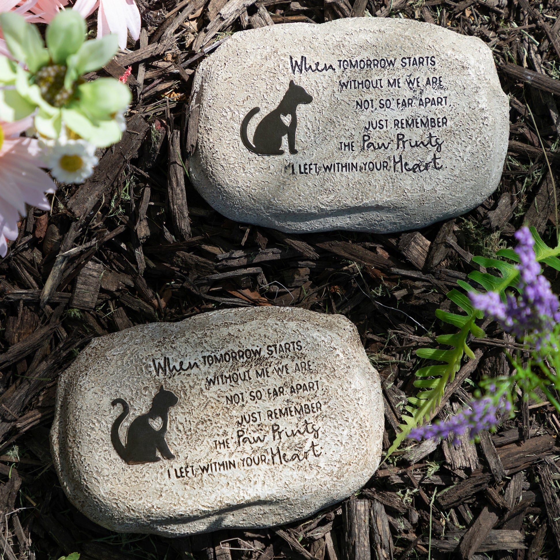 Two engraved stones with cat and dog illustrations and text on a mulched surface with flowers.