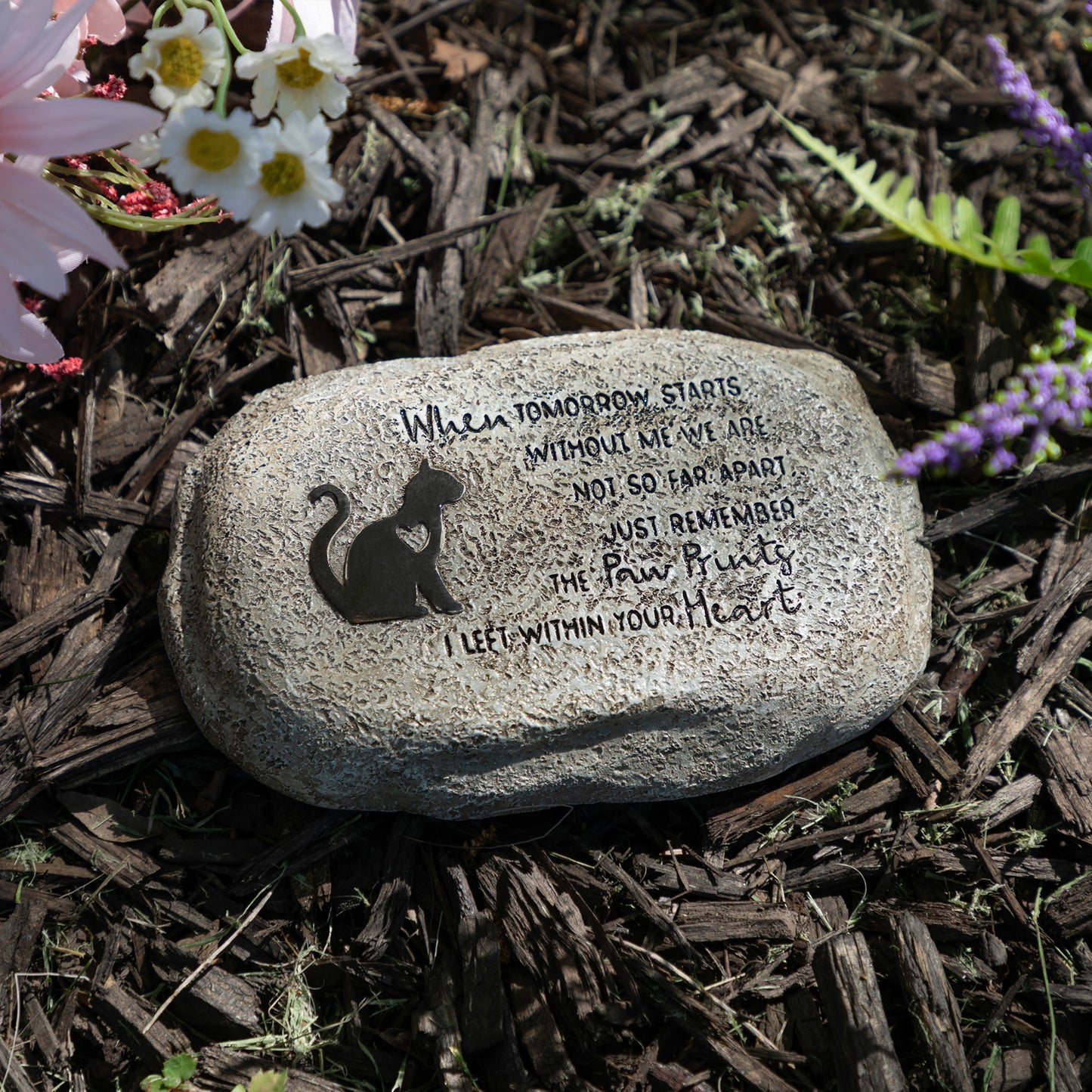 Decorative stone with a cat silhouette and engraved text on a mulched garden bed with flowers.