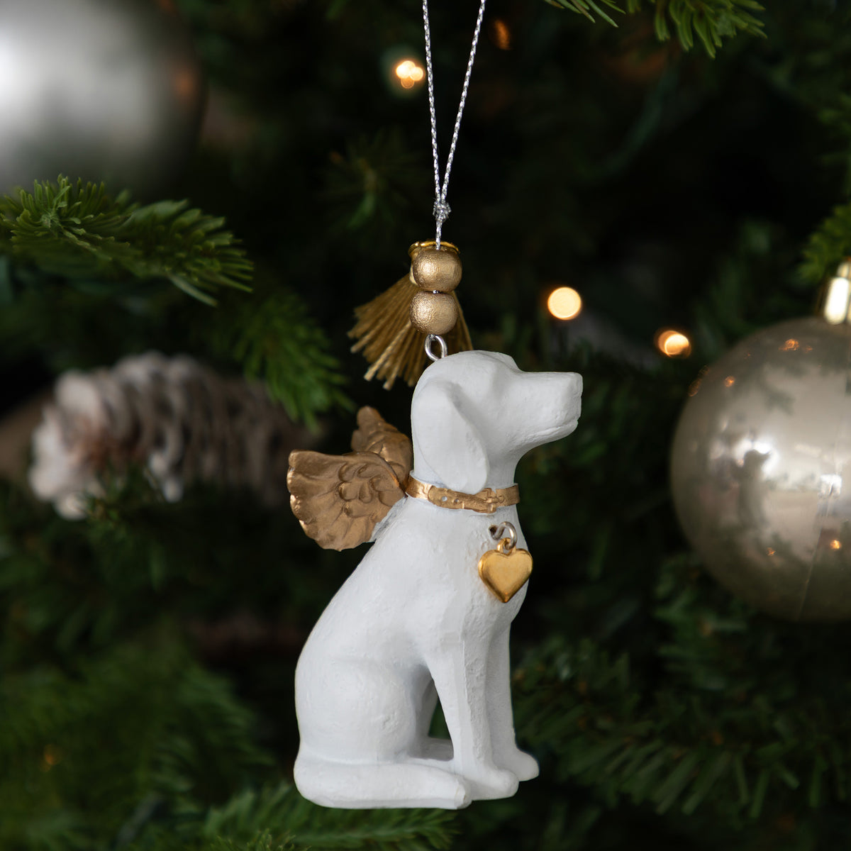 A white dog-shaped Christmas ornament with gold angel wings, a gold collar, and a gold heart-shaped tag, hanging on a decorated Christmas tree.