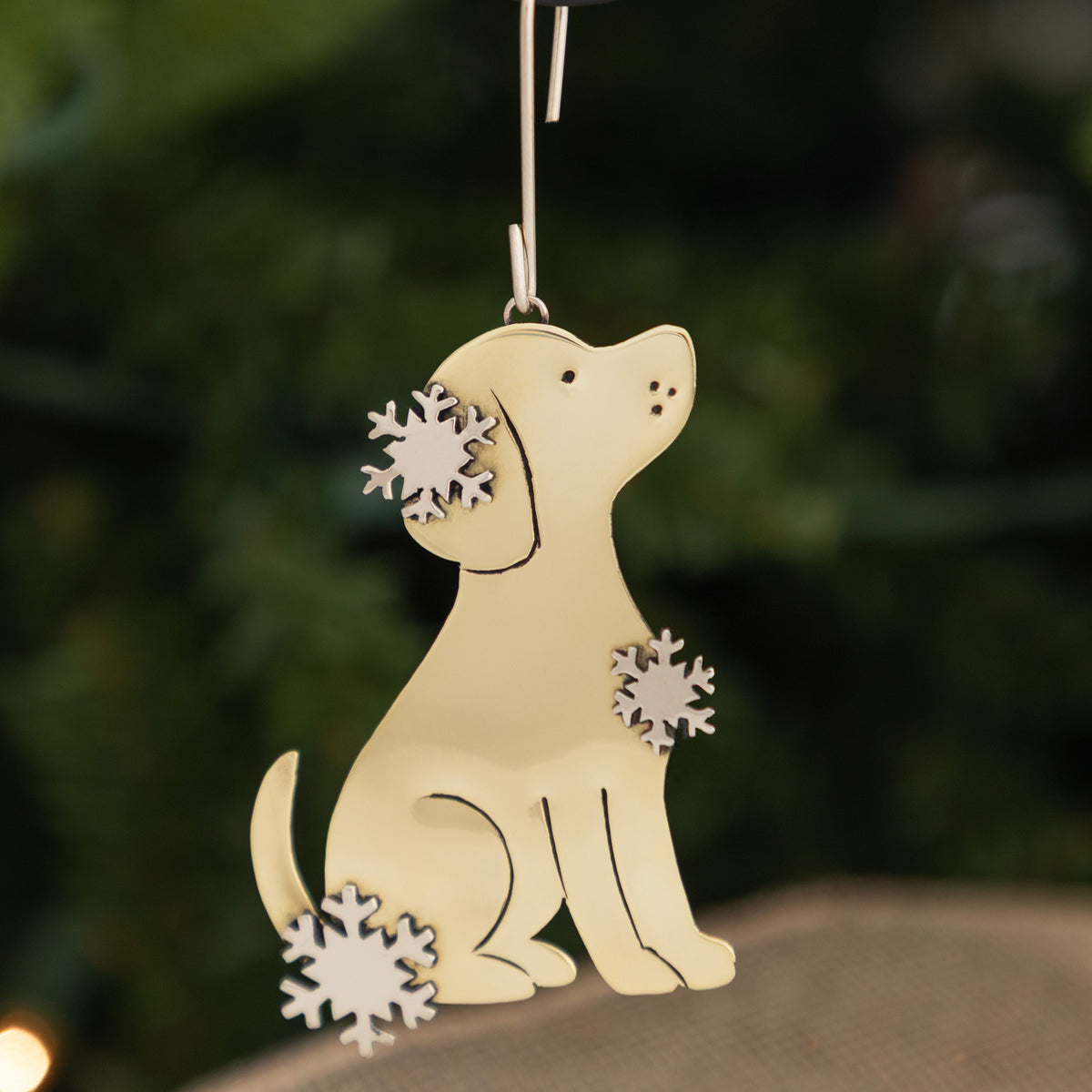 A hanging Christmas ornament shaped like a sitting dog in pale yellow, decorated with three white snowflake cutouts, photographed against a blurred green outdoor background.