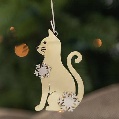 A light cream-colored metal cat Christmas ornament with silver snowflake accents, hanging against a dark green blurred background with warm bokeh lights.