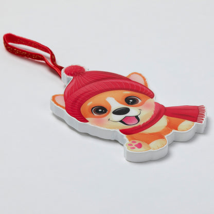 A cartoon dog ornament featuring a smiling orange and cream dog wearing a red hat and red scarf, with a red hanging loop, photographed on a plain light background.