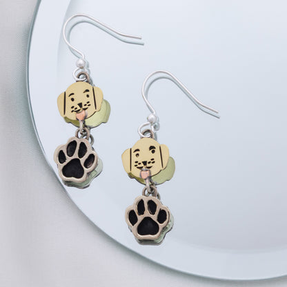 A pair of dangling dog-themed earrings resting on a mirrored surface. Each earring has a silver fishhook wire, a small brass charm shaped like a dog's head with black facial details, and a dangling paw print charm in silver with a black center.