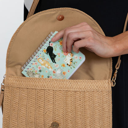 A tan woven crossbody bag with a hand placing a small floral notebook with cat illustrations into its open front pocket.
