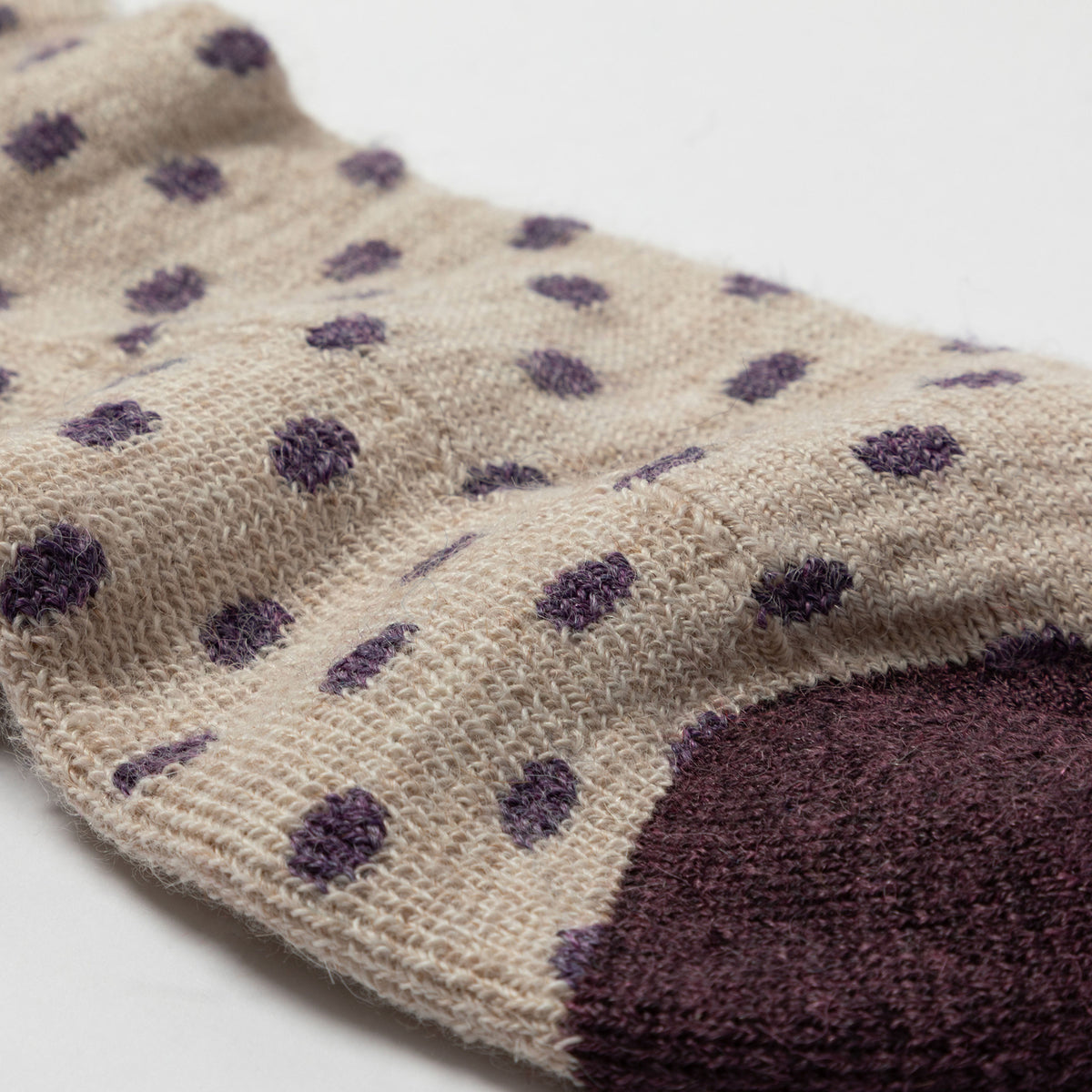 A close-up of a knit sock in a warm beige color with scattered dark purple polka dots and a solid dark purple toe section, shown on a plain white background.