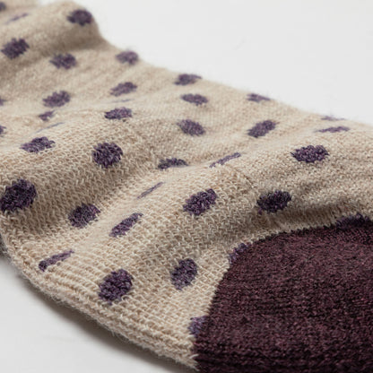 A close-up of a knit sock in a warm beige color with scattered dark purple polka dots and a solid dark purple toe section, shown on a plain white background.