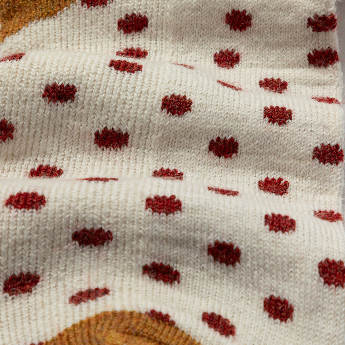 Close-up of a knitted fabric with a cream base and scattered dark red polka dots, showing the knit texture and part of a tan circular pattern at the bottom edge.