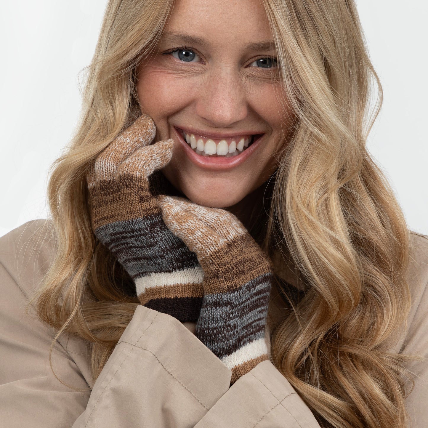 A smiling blonde woman wearing a pair of knit striped gloves in brown, beige, gray, and white, paired with a beige coat against a plain light background.