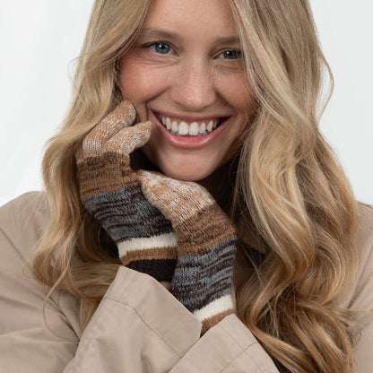 A smiling blonde woman wearing a pair of knit striped gloves in brown, beige, gray, and white, paired with a beige coat against a plain light background.