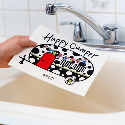 Hand holding a 'Happy Camper' towel over a sink with a faucet.