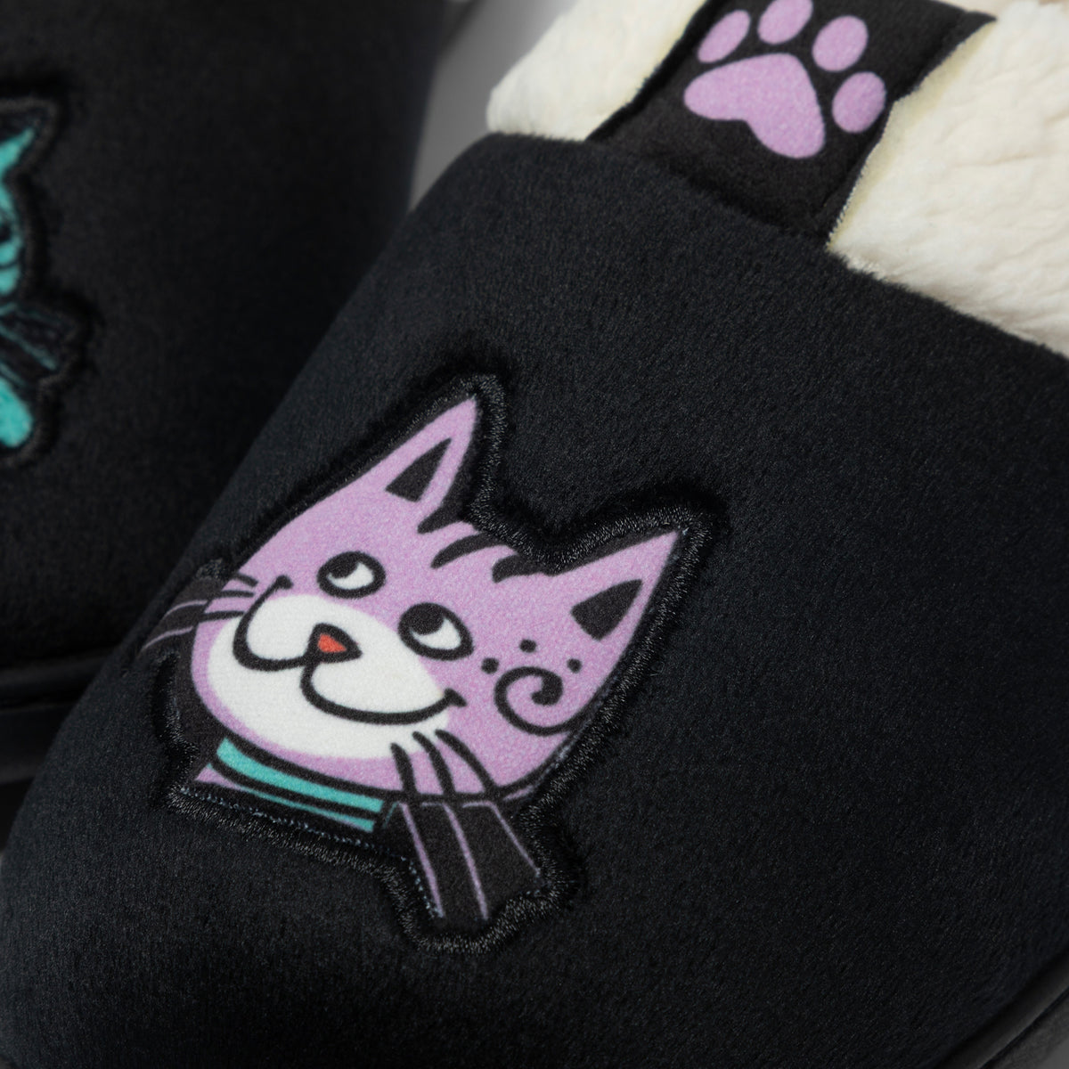 Close-up of a black plush slipper with a large embroidered purple cat face patch on the side and a white sherpa lining visible at the opening.