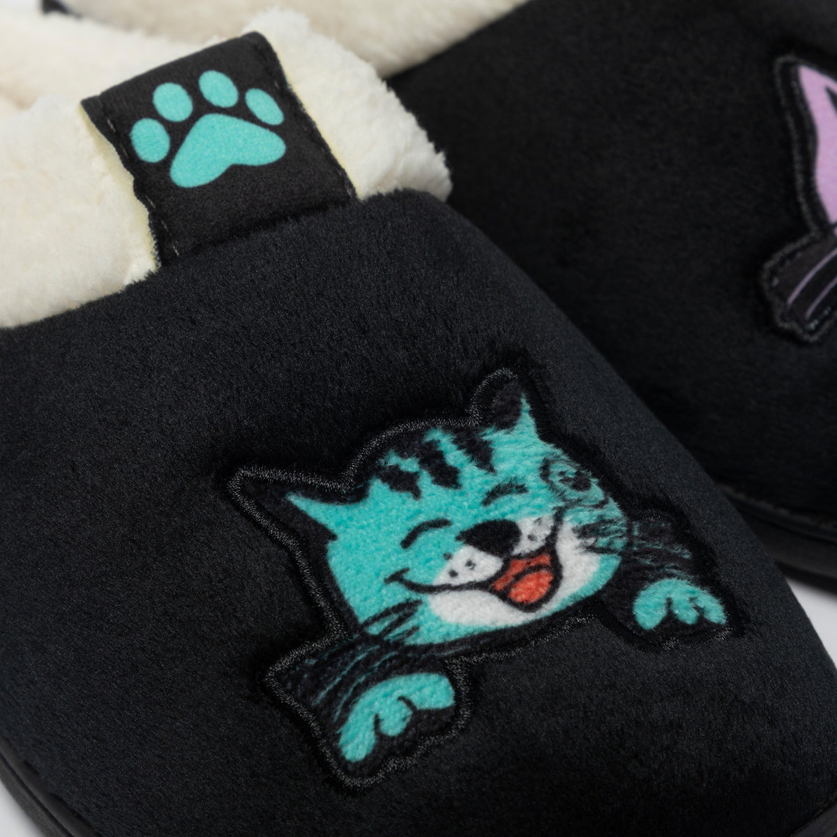 Close-up of a pair of black plush slippers with white sherpa lining. The front slipper features a large embroidered teal cat face with a red tongue and black outlines, and a matching teal paw print patch on the tab.