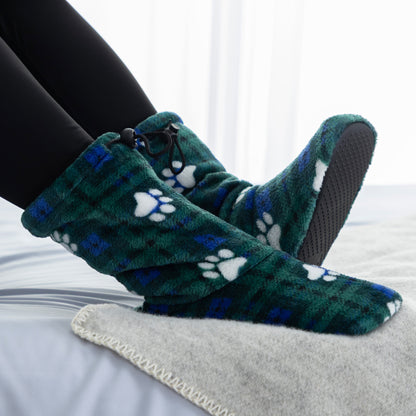 A person wearing plush slip-on slippers with a green plaid pattern and white paw prints, standing on a light gray blanket on a bed.