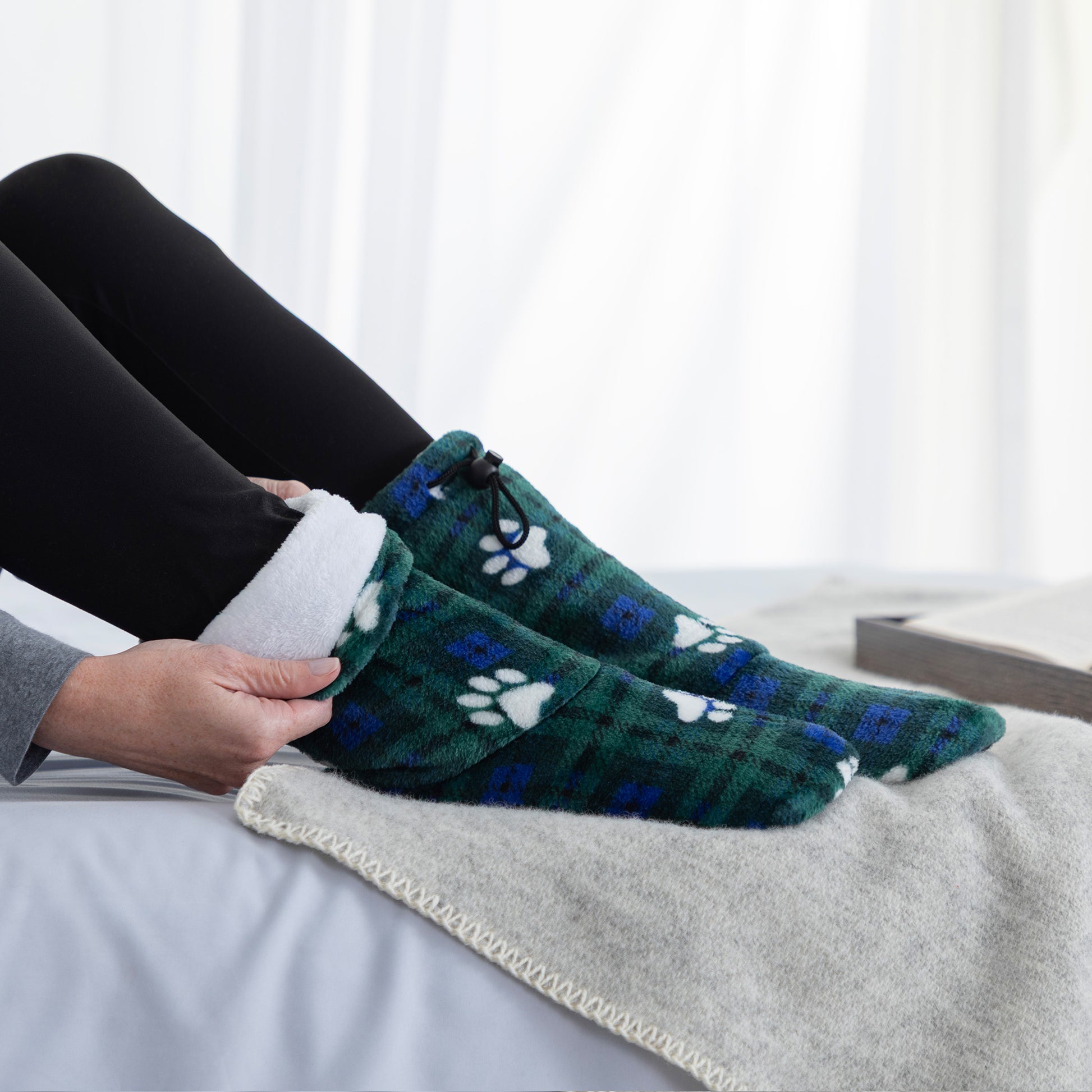 A person wearing green plaid socks with white paw prints, shown sitting on a bed with a blanket. The socks have a soft fleece-like texture and a white fuzzy lining.