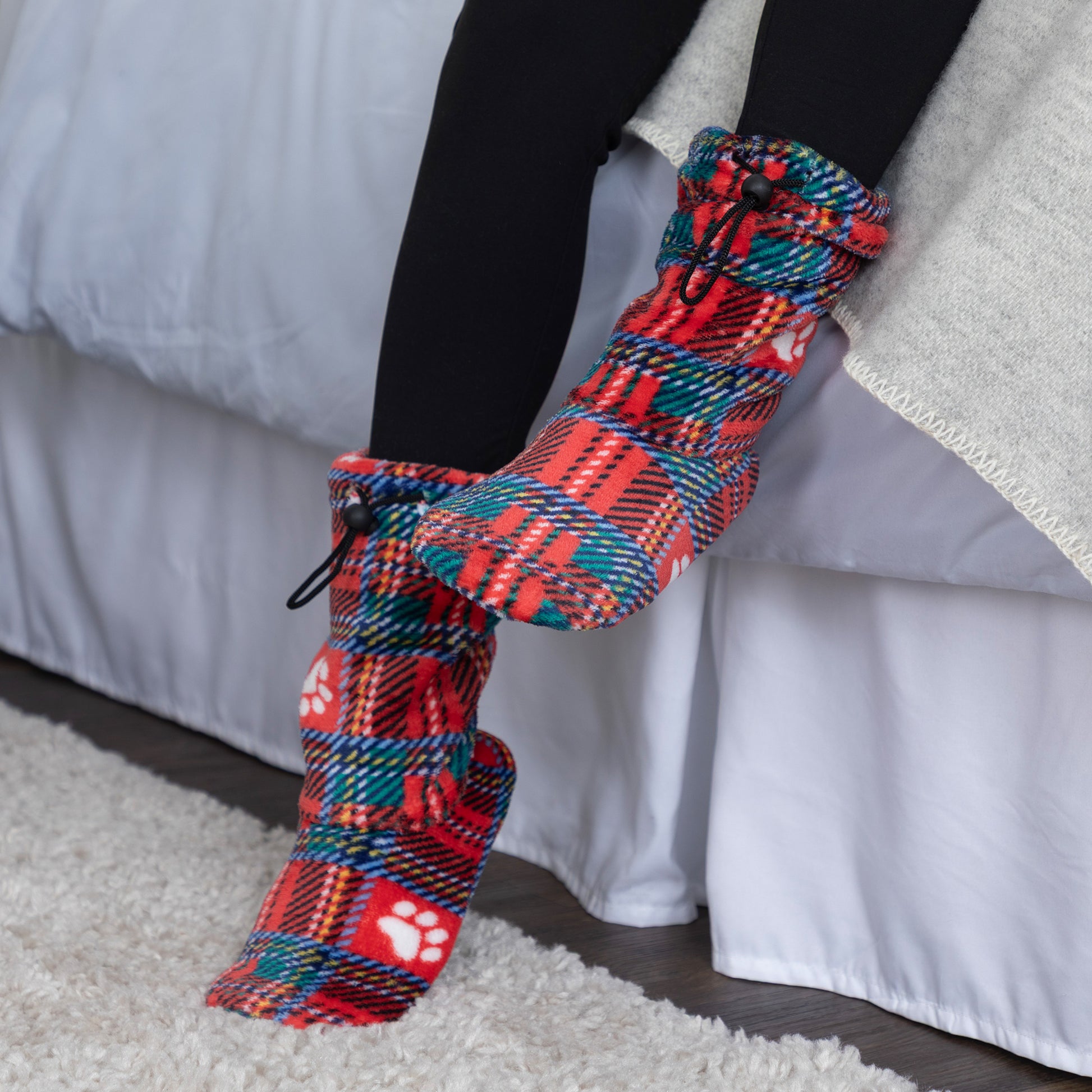 A person wearing a pair of colorful knit booties or clogs with a red, teal, and black plaid pattern, white paw prints, and a black drawstring, shown sitting on a bed in a bedroom setting.