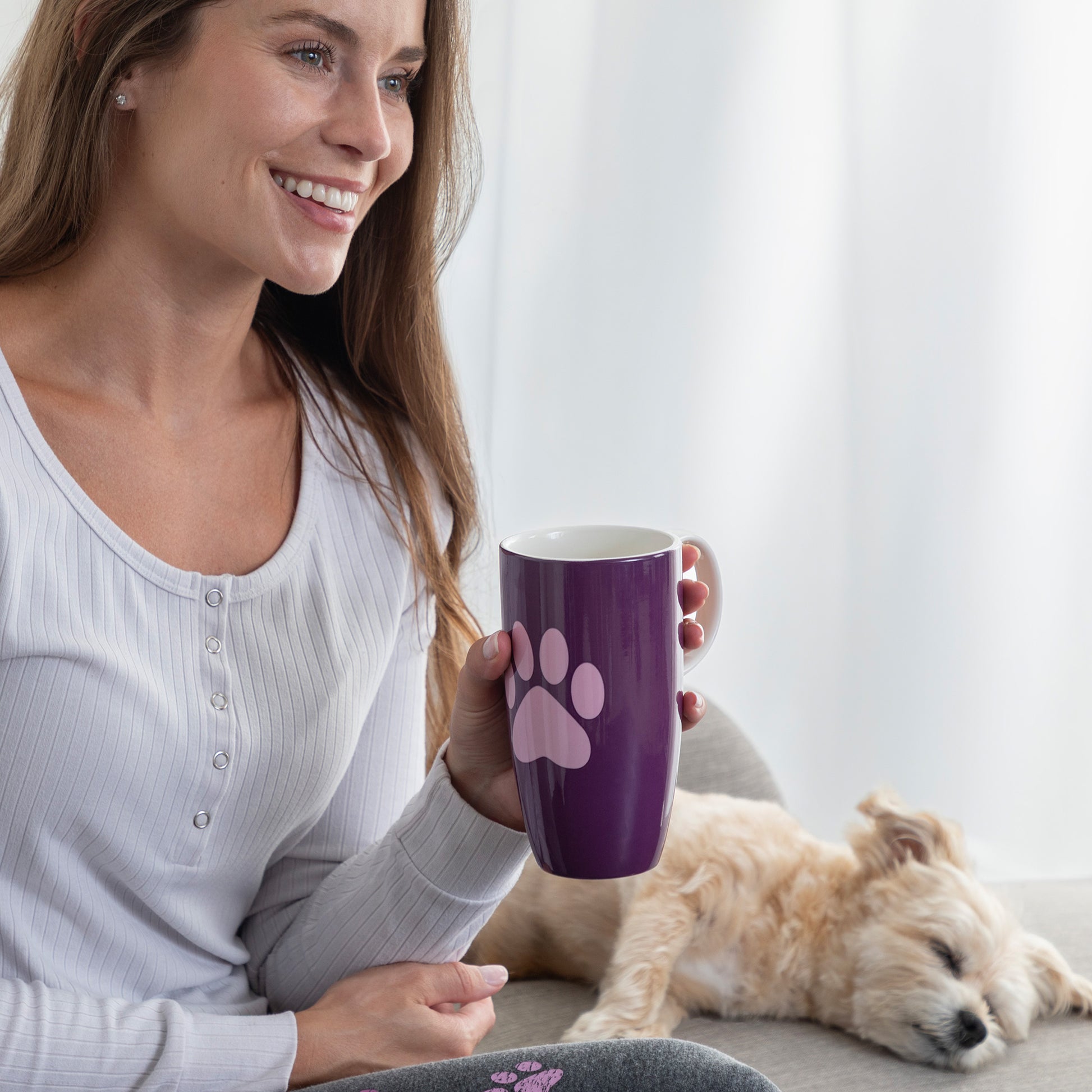 A woman in a white ribbed henley shirt holds a purple ceramic mug with a pink paw print design. A small light-colored dog is sleeping on a couch in the background.