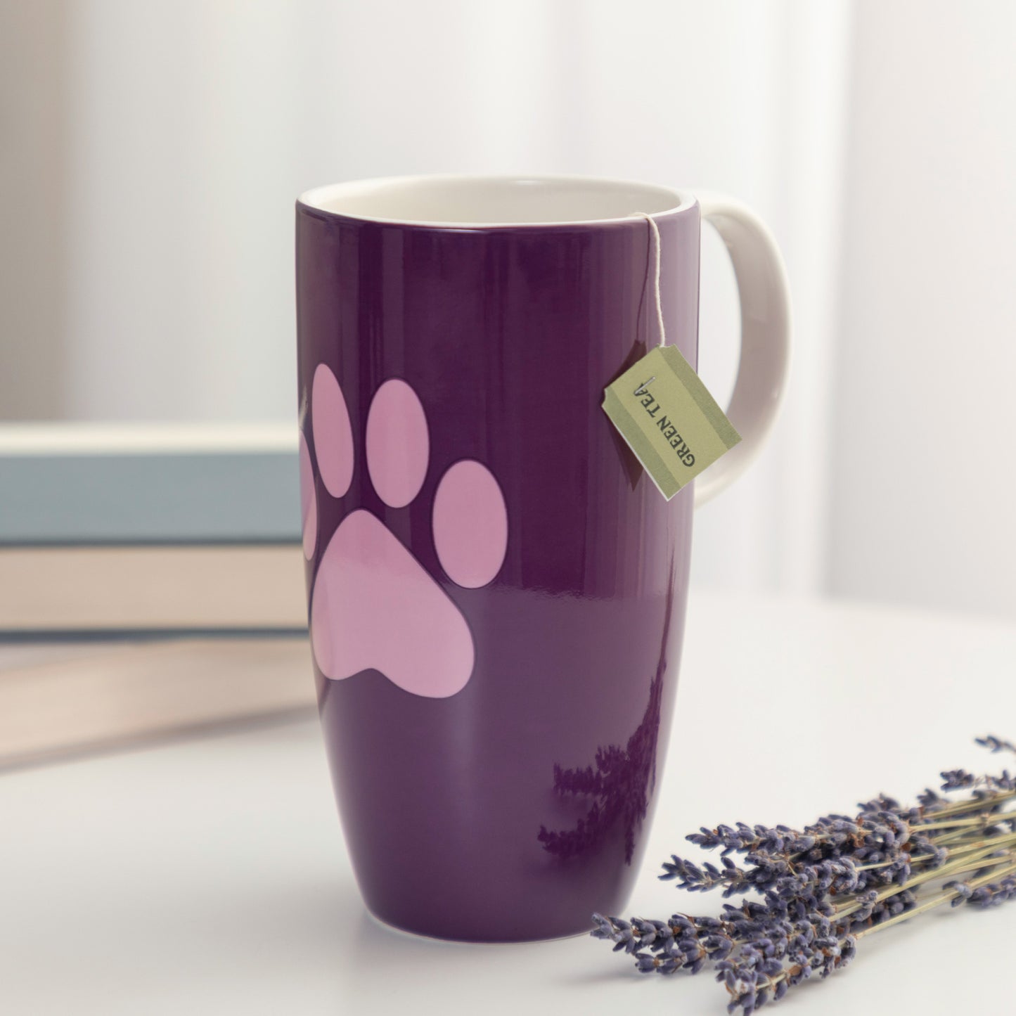 A purple ceramic mug with a purple paw print design, featuring a tea bag tag, set on a white surface with lavender and books in the background.