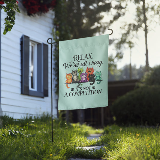 A light mint green garden flag with a humorous cat slogan and a row of colorful cartoon cats, displayed on a black metal stand in a grassy yard in front of a white house.