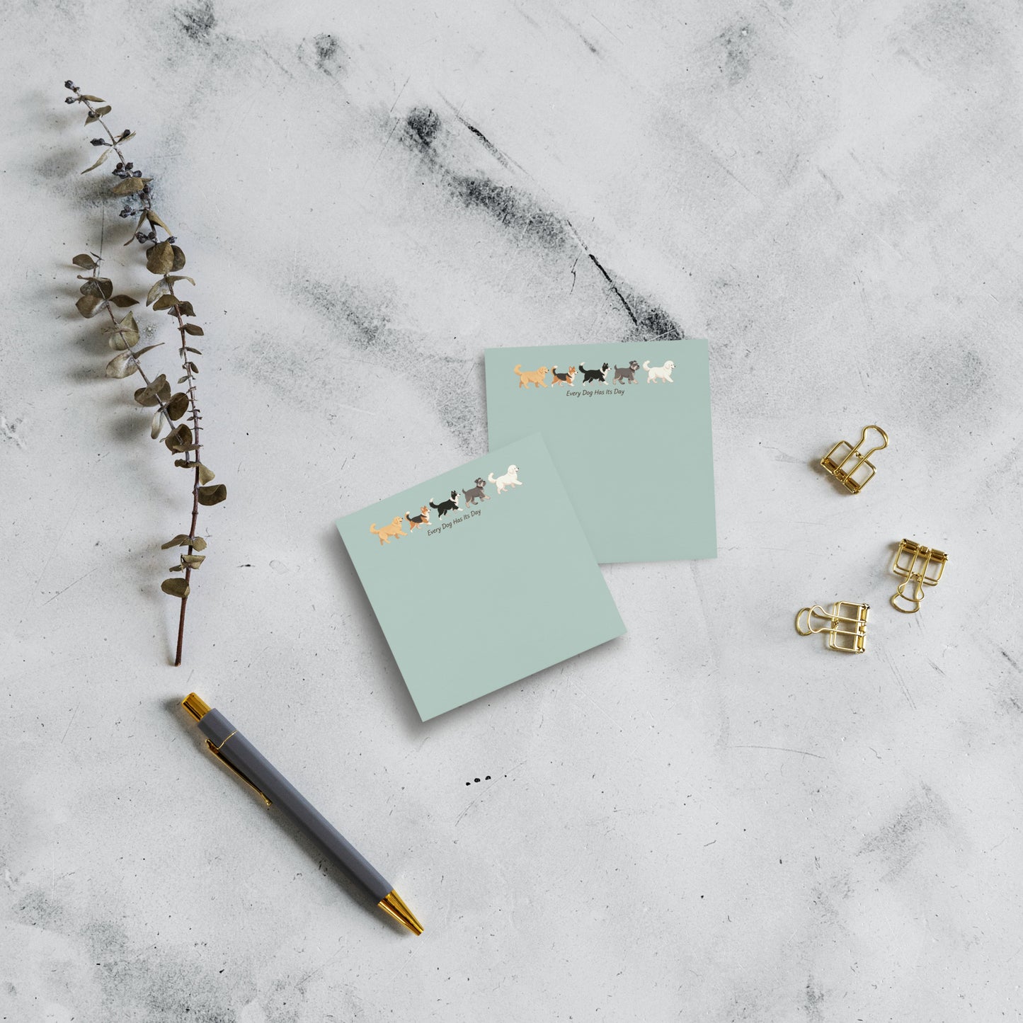 Two 3x3 mint green notepads with a cute dog illustration and the text 'Every Dog has Its Day' at the top, styled on a light gray marble background with a gray pen, gold paperclips, and a dried eucalyptus branch.