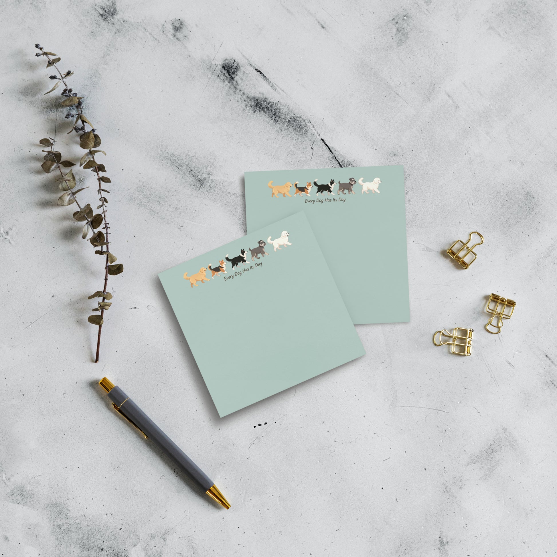 Two 4x4 square mint green sticky notes with a row of colorful dog illustrations and the text 'Every Dog Has Its Day' are arranged on a light gray marble surface with a gray pen, gold binder clips, and a dried eucalyptus branch.