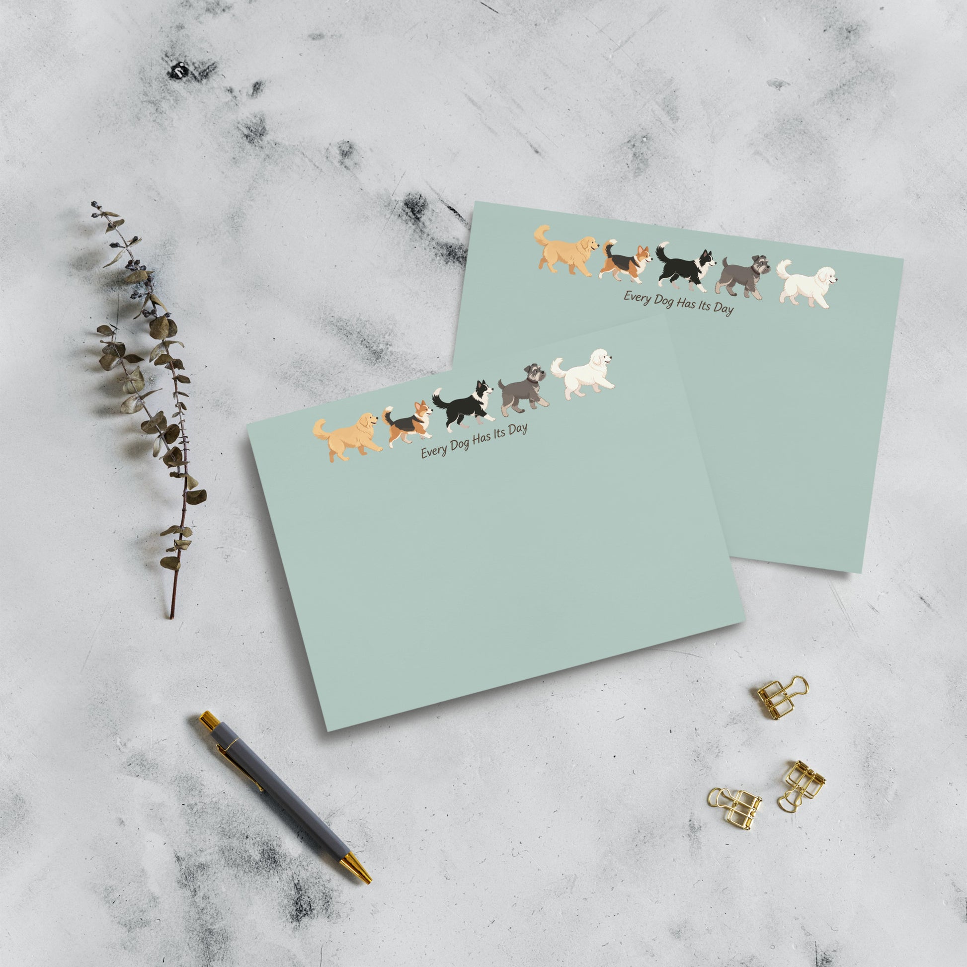 Two 8x6 mint green notepads with a row of colorful dogs and the text 'Every Dog Has its day' are arranged on a light marble surface with a pen, paper clips, and a sprig of eucalyptus.