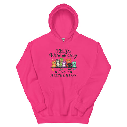 A bright pink pullover hoodie with a graphic on the chest featuring the text 'RELAX, We're all crazy' and 'IT'S NOT A COMPETITION' alongside a row of colorful cartoon cats in different poses.