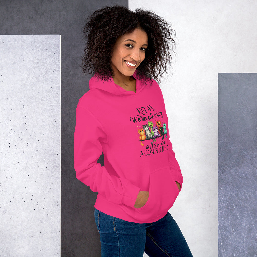A woman wearing a bright pink hoodie with a graphic that reads 'RELAX We're all crazy it's not A COMPETITION' with cartoon animals, paired with blue jeans, set against a split gray and white textured background.