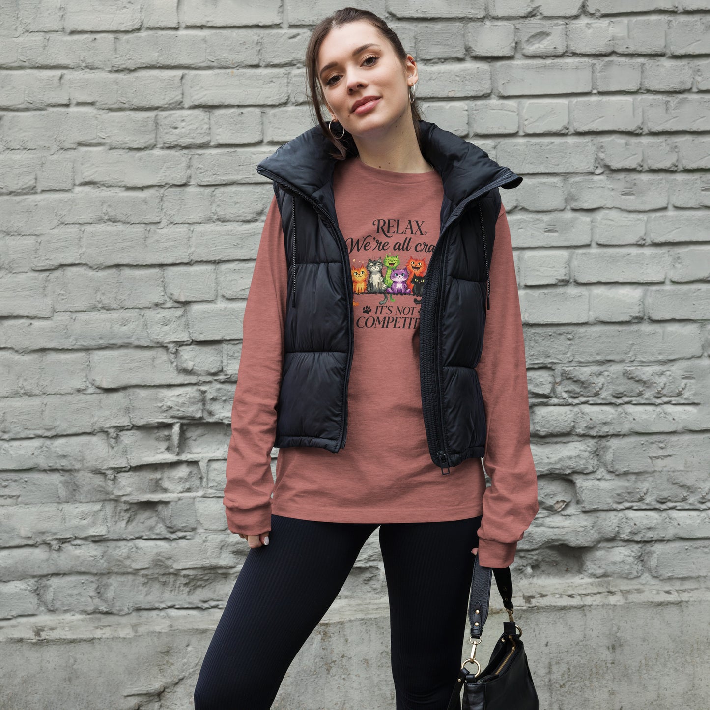 A woman stands against a gray brick wall wearing a black puffer vest over a dusty rose graphic long-sleeve shirt and black leggings, holding a black handbag.